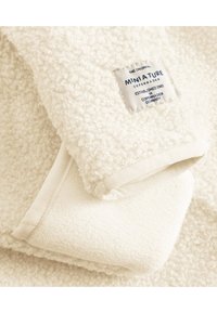 Cremefarbene Fleece-Decke mit einer strukturierten, geschlungenen Oberfläche. Verfügt über einen weichen, glatten Rand und ein Markenetikett in der Ecke.