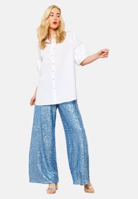 Camicione bianco oversize con maniche corte e plissettate, abbinato a pantaloni larghi blu in paillettes, che mostrano una texture brillante e riflettente.