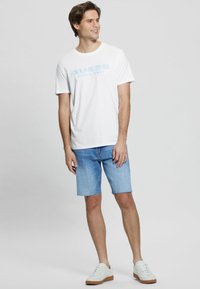 Witte katoenen T-shirt met blauwe geprinte "GUESS" logo, gecombineerd met lichtblauwe denim shorts en witte sneakers met tan kleurige zolen.
