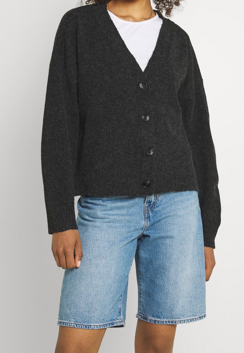 Cárdigan de punto gris oscuro con escote en V, mangas largas y cinco botones negros, combinado con pantalones cortos de mezclilla azul claro. Textura suave.
