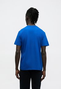 Niebieska bawełniana koszulka z krótkim rękawem, okrągłym dekoltem i gładką fakturą, widziana od tyłu. Brak widocznych wzorów lub elementów metalowych.