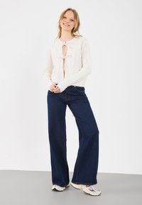 Kremfarget kabelstrikket genser med bindefront, parret med mørkeblå vide jeans og multifargede joggesko. Ren, enkel design.