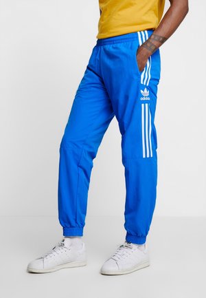 Mann trägt blaue Adidas-Trainingshose mit weißen Streifen und weißen Sneakers, steht mit einer Hand in der Tasche.