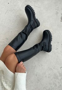 Zwarte kniehoge laarzen van glad synthetisch materiaal, met een stevige zool, gestructureerde accenten en een zijrits voor eenvoudig aan- en uittrekken.