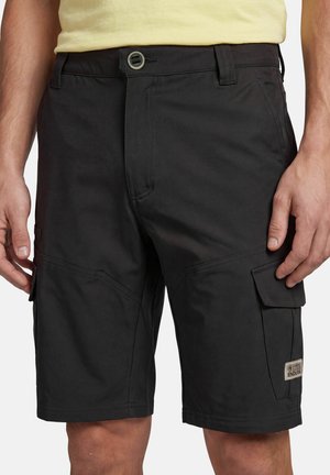 Schwarze Cargoshorts mit Front- und Seitenklappentaschen, getragen von einer stehenden Person in einem gelben Hemd, von der Taille bis zu den Knien sichtbar.