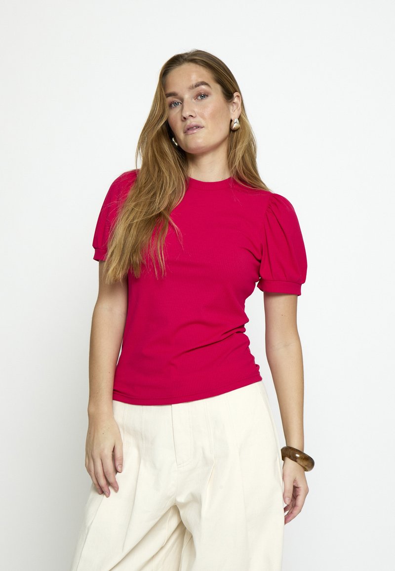 Frau mit langen hellbraunen Haaren trägt ein leuchtend pinkfarbenes Oberteil mit Puffärmeln, cremefarbene weite Hosen, goldene Ohrringe und ein dunkles Armband, sie steht vor einem weißen Hintergrund.