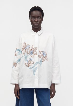 Femme portant une chemise blanche boutonnée avec une broderie florale bleue et marron sur le devant et les manches, associée à un pantalon bleu à jambes larges.
