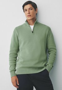 Groene trui met rits en een gestructureerd breipatroon, met geribbelde mouwen en onderkant. Gekozen boven een wit T-shirt en gecombineerd met donkere broek.