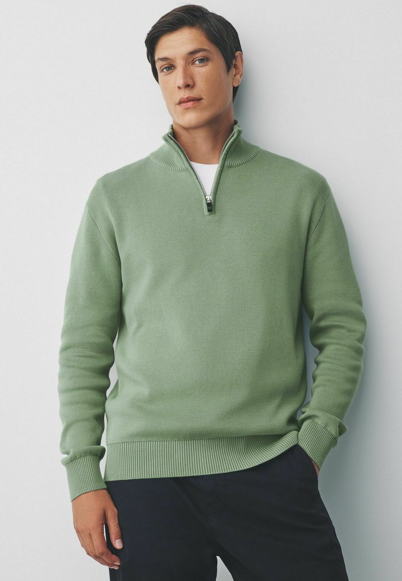 Groene trui met rits en een gestructureerd breipatroon, met geribbelde mouwen en onderkant. Gekozen boven een wit T-shirt en gecombineerd met donkere broek.