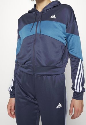 Personne portant un survêtement Adidas bleu marine et bleu sarcelle avec un sweat à capuche zippé et un pantalon assorti avec des bandes blanches et des logos.