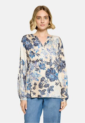 Kvinde iført en beige bluse med lange ærmer med blå blomsterprint og blå jeans, stående mod en ensfarvet lys baggrund.