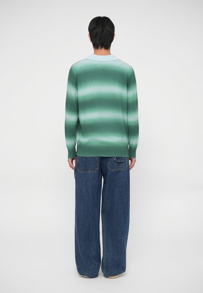 Maglione verde a gradiente con maniche lunghe, polsini a coste e colletto azzurro chiaro. Indossato con jeans scuri a taglio morbido.
