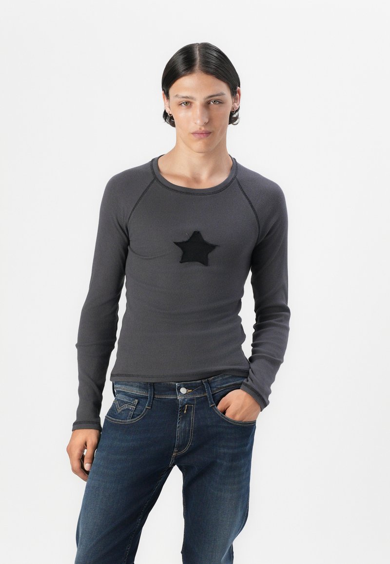 Graues Langarmshirt aus geripptem Stoff mit einem schwarzen Sternaufnäher auf der Brust, kombiniert mit blauen Jeans.