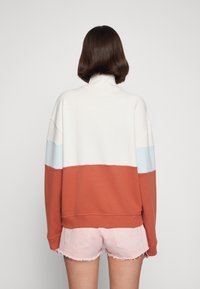 Sweatshirt i krämfärg, rostfärg och ljusblå, med hög krage och lös passform. Mjuk textur och ribbade ärmslut. Matchas med rosa shorts.
