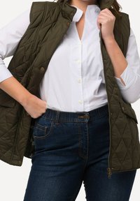 Frau in einer dunkelgrünen gesteppte Weste über einem weißen Hemd und dunkelblauen Jeans, mit einer Hand in der Tasche und der anderen, die den Weste-Kragen hält.