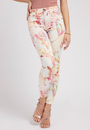 Femme portant un pantalon slim blanc à imprimé floral rose et orange, associé à des talons à brides en or rose et un top rose sans manches.