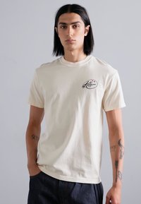 Cream cotton T-shirt with short sleeves, round neckline, and a small embroidered logo on the left chest. Model stands against a gray background.