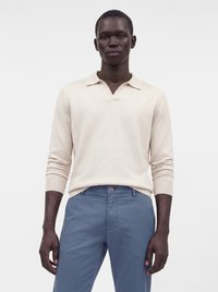 Homme portant un pull polo beige clair à manches longues et un pantalon bleu, debout devant un fond blanc uni.