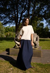 Cream knitted short-sleeve top and long navy skirt, earthy texture with a smooth finish. Background includes greenery and a stone sculpture.