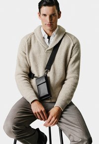 Beige ribbed sweater with a shawl collar, worn over a white shirt. Black crossbody bag rests against a grey patterned pant.