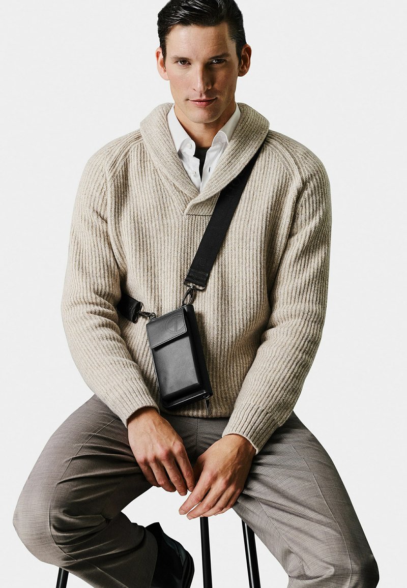 Beige ribbed sweater with a shawl collar, worn over a white shirt. Black crossbody bag rests against a grey patterned pant.