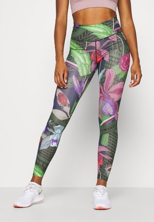 Personne athlétique portant des leggings taille haute avec des motifs floraux et de feuilles colorés, assortis à des baskets blanches et un crop top rose clair.