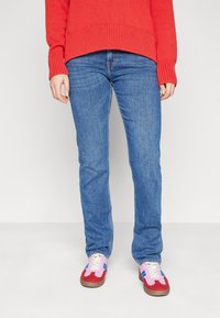 Červený žebrovaný svetr, modré slim-fit džíny a vícbarevné tenisky s růžovými a červenými akcenty, které mají bílé tkaničky a modrý pruh.