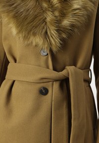 Oliven grøn frakke med faux fur-krave, frontknapper og bånd i taljen. Glat stoftekstur med en flatterende silhuet.