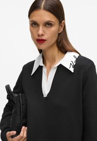Sudadera negra con cuello blanco que presenta texto bordado. El modelo sostiene un bolso de mano negro. Tejido suave con un corte relajado y diseño de cuello en V.