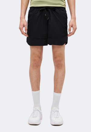 Persona che indossa pantaloncini neri con coulisse, calzini bianchi alti e scarpe da ginnastica bianche, in piedi davanti a uno sfondo chiaro e uniforme.