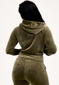 Olívazöld velúr melegítő, kapucnis dzsekivel és hozzáillő nadrággal. A hátán nagy, köves "Juicy" felirat látható.