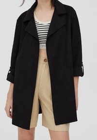 Chaqueta negra de corte holgado hecha de un tejido suave, con un amplio cuello, mangas enrolladas y bolsillos laterales, combinada con una camiseta a rayas y pantalones cortos beige.