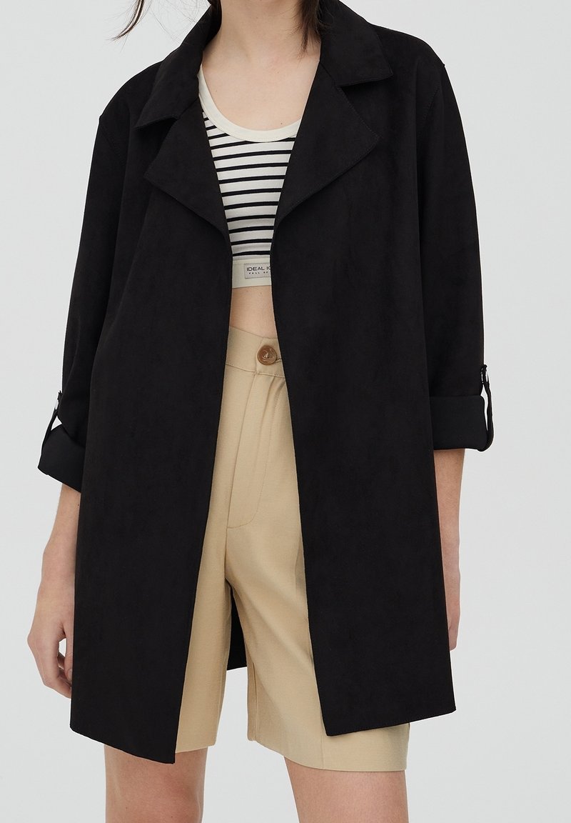 Chaqueta negra de corte holgado hecha de un tejido suave, con un amplio cuello, mangas enrolladas y bolsillos laterales, combinada con una camiseta a rayas y pantalones cortos beige.