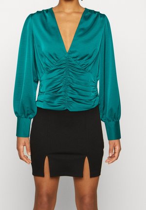 Blusa di raso color teal con maniche a sbuffo lunghe e scollatura a V profonda; presenta arricciature in vita. Abbinata a una mini gonna nera con spacco.