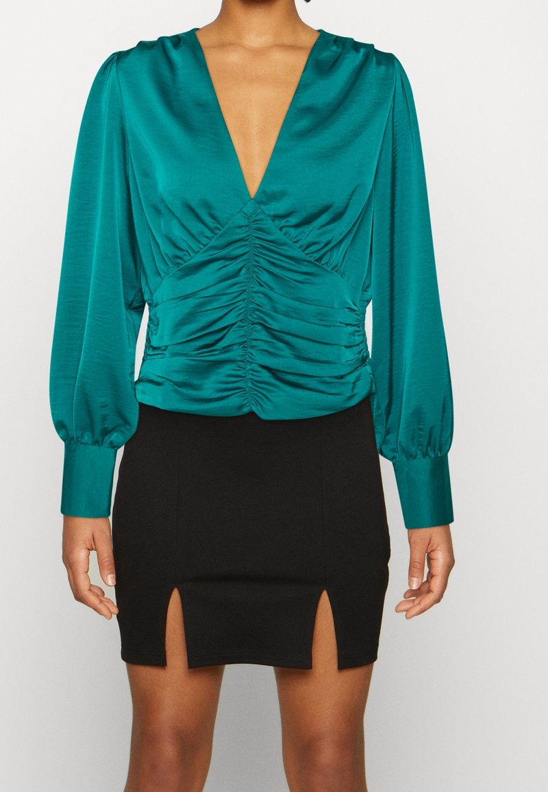 Blusa di raso color teal con maniche a sbuffo lunghe e scollatura a V profonda; presenta arricciature in vita. Abbinata a una mini gonna nera con spacco.