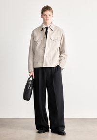 Világosszürke szarvasbőr dzseki két mellzsebbel, bézs ing, fekete nyakkendő és fekete, bőszárú nadrág. Fekete táska ívelt fogantyúkkal.