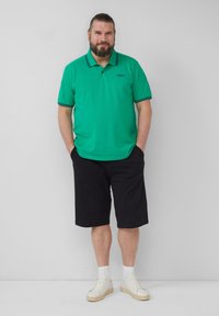 Groene poloshirt met zwarte accenten, gecombineerd met zwarte shorts. De outfit omvat witte sneakers en een effen achtergrond.