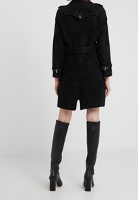 Vrouw in een zwarte suède trenchcoat met riem en zwarte leren kniehoogte hakken, staande op een witte naadloze achtergrond.
