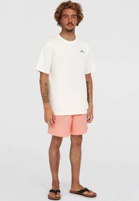 Bărbat cu păr creț, purtând un tricou alb O'Neill, pantaloni scurți roz, șlapi negri și cu tatuaje vizibile pe brațe, stând în fața unui fundal neutru.