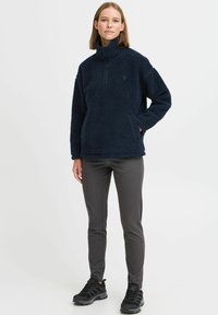 Marineblå fleecegenser med trakt hals, halv glidelås og teksturert overflate. Kombinert med grå slim-fit bukser og svarte joggesko.