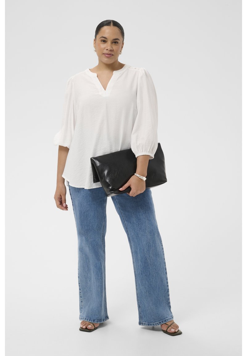 Femme en blouse blanche et jean bleu tenant une grande pochette noire, portant des sandales ouvertes et un bracelet argenté, debout devant un fond blanc.