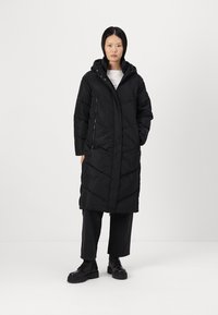 Cappotto lungo imbottito nero con cappuccio, caratterizzato da un motivo geometrico a trapunta, zip frontale e tasche laterali. Indossato sopra pantaloni neri e stivali.