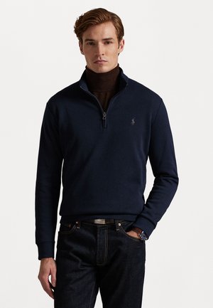 Pull-over bleu marine à mi-fermeture éclair en tissu doux, featuring un col et des poignets côtelés, avec un petit logo brodé sur la poitrine gauche. Associé à un jean foncé.