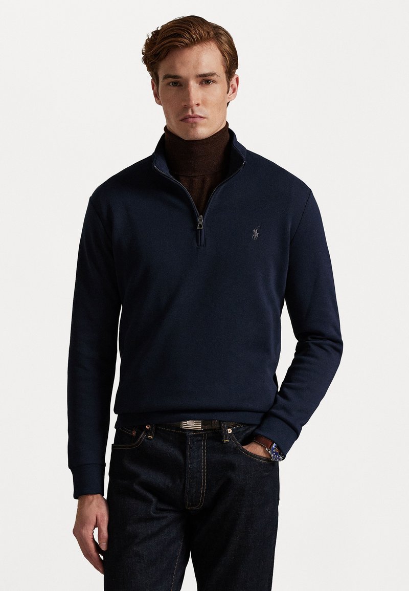 Mørkeblå half-zip genser i myk stoff, med en ribbestrikket krage og mansjetter, samt en liten brodert logo på venstre bryst. Kombinert med mørke denimbukser.