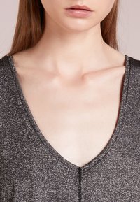 Top senza maniche, nero e luccicante, con un design a scollatura profonda a V. Il materiale ha una finitura metallica; la cucitura piatta lungo il colletto esalta l'estetica moderna.
