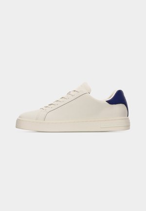White low-top sneaker with navy heel tab, lace-up closure, and flat sole shown in side profile on neutral background.