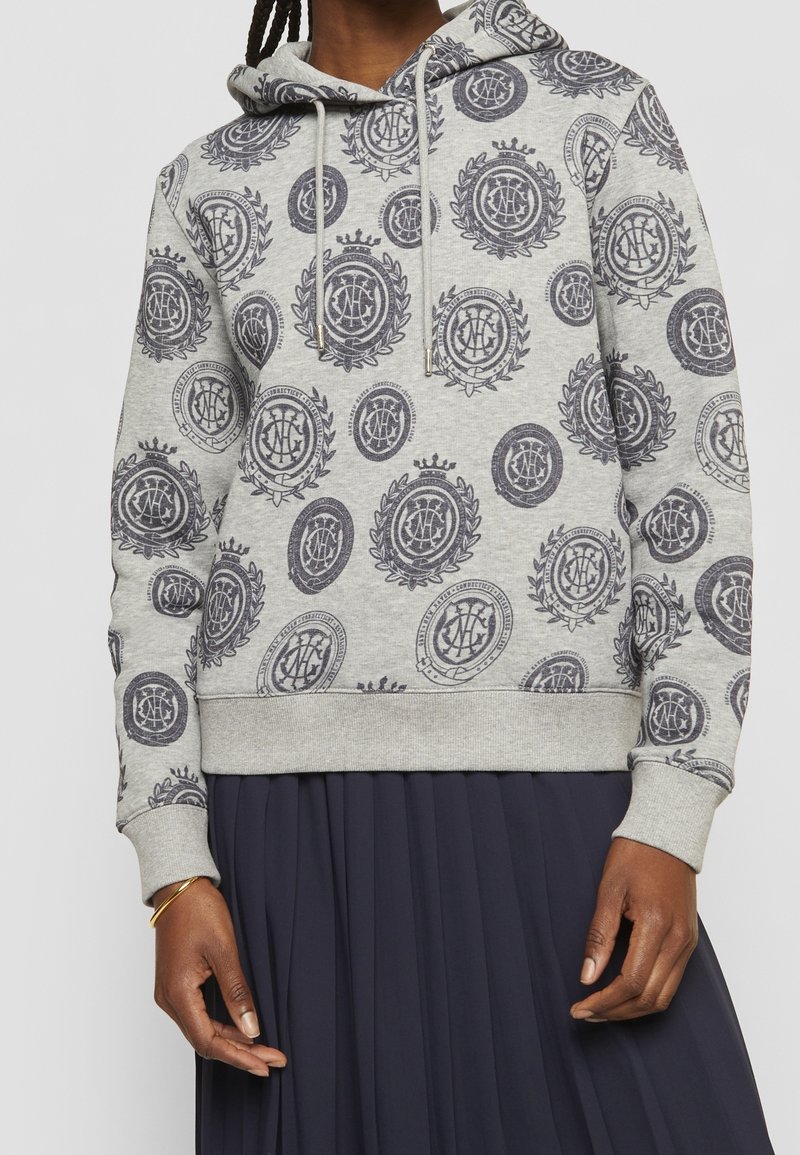 Personne portant un sweat à capuche gris clair avec des motifs circulaires foncés et une jupe plissée bleu marine, debout avec les mains détendues visibles.