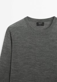 Pull en laine gris avec un col rond. Présente une texture lisse et un style minimaliste. L'étiquette indique "100% laine" et le nom de la marque.