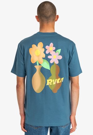 Osoba z krótkimi włosami w niebieskiej koszulce, na której z przodu znajdują się dwa stylizowane wazy trzymające żółte i różowe kwiaty, a z tyłu napis "RVCA".
