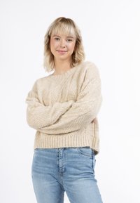 Beige kötött pulóver bordás szegéllyel és textúrált mintával, világoskék farmernadrággal párosítva. Az anyag lágy és meleg hatású.